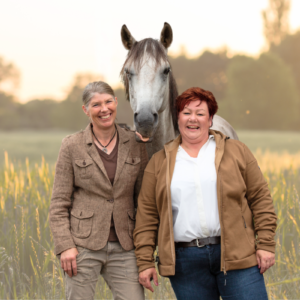 Zwei Frauen stehen lächelnd in die Herbstsonne mit einem Pferd auf der Wiese.