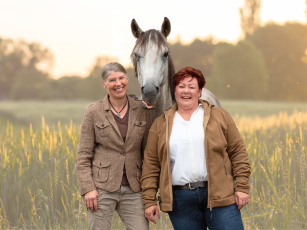 Zwei Frauen stehen lächelnd in die Herbstsonne mit einem Pferd auf der Wiese.