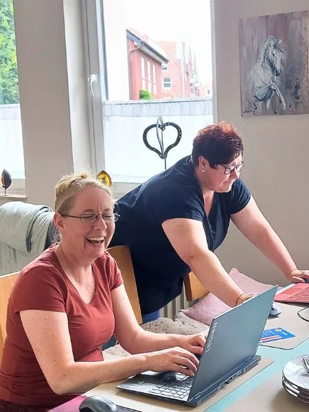 Zwei Frauen sitzen mit einem Laptop lachend an einem Tisch.