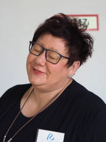 Eine Frau mit kurzen Haaren und Brille, die verschmitzt Augen und Mund zukneift.