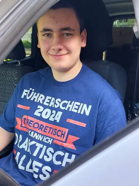 Ein junger Mann sitzt mit blauem T-Shirt in einem weißen Auto.
