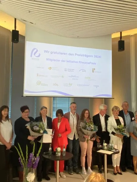 Eine große Menschengruppe hält Blumen und steht vor einer Leinwand.