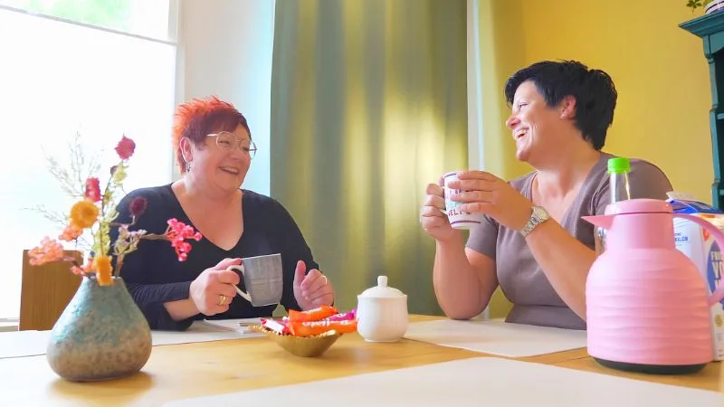 Zwei Frauen sitzen am Tisch, halten Tassen und reden.