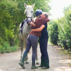 Angstfrei Reiten: Zwei Frauen stehen im Reitercoaching auf einem Waldweg neben einem Pferd und umarmen sich.