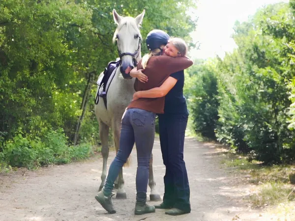 Angstfrei Reiten: Zwei Frauen stehen im Reitercoaching auf einem Waldweg neben einem Pferd und umarmen sich.