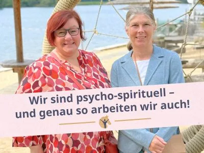 Blockgrafik: „Wir sind psycho-spirituell - und genau so arbeiten wir auch!“; Jutta und Denise Beckmann stehen vor einem See und lächeln in die Kamera.