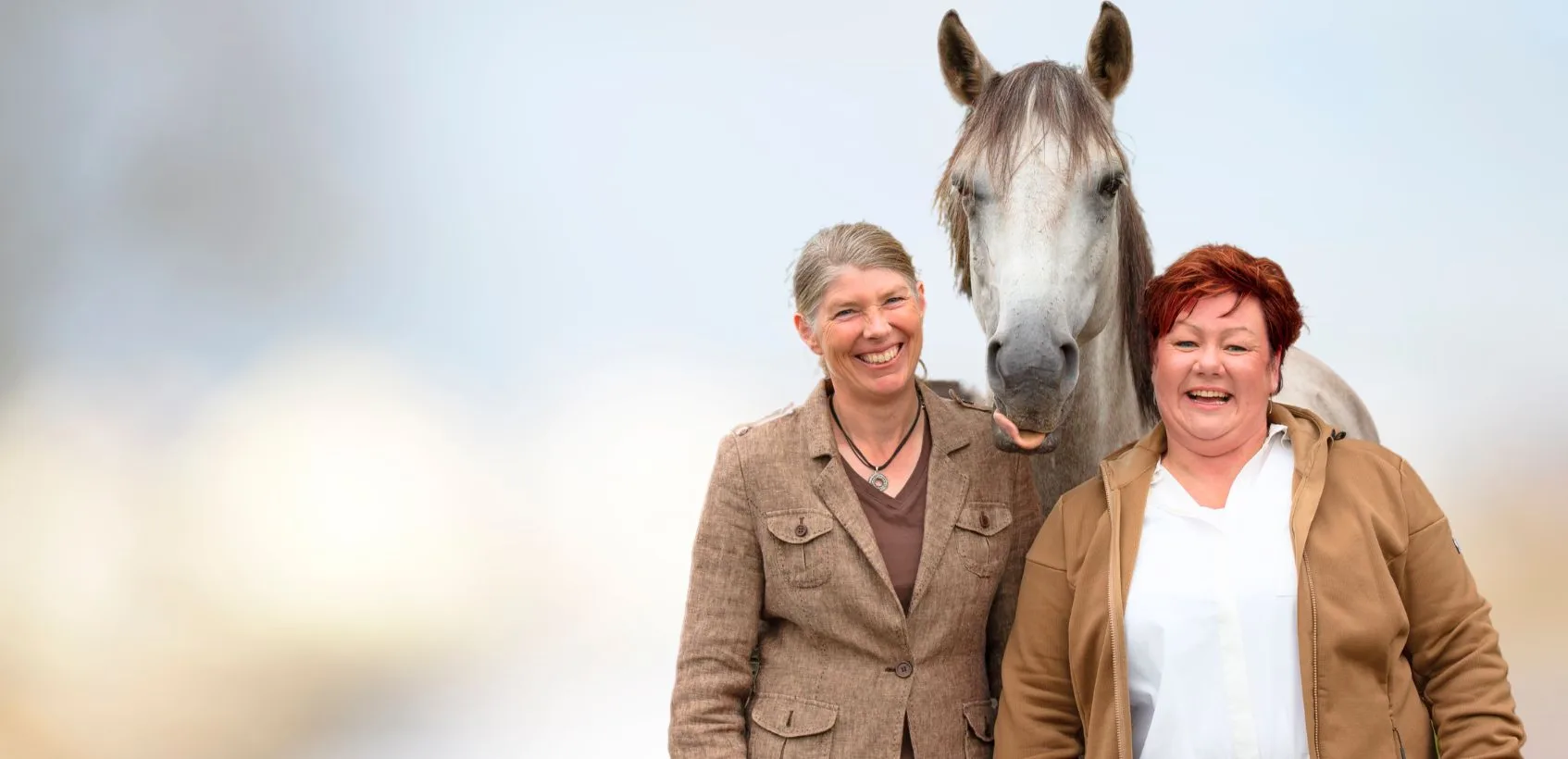 Zwei Frauen, hinter denen ein Pferd steht und die Zunge rausstreckt.