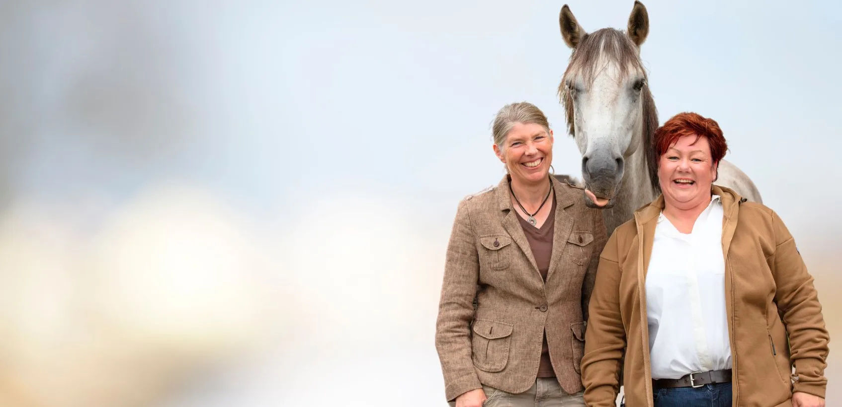 Jutta und Denise Beckmann stehen vor einem weißen Pferd, das die Zunge rausstreckt.