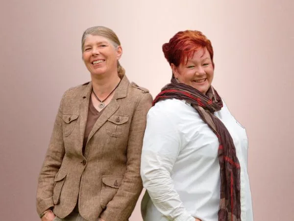 Jutta und Denise Beckmann stehen Rücken an Rücken, sind zur Kamera gewandt und schauen dich an.